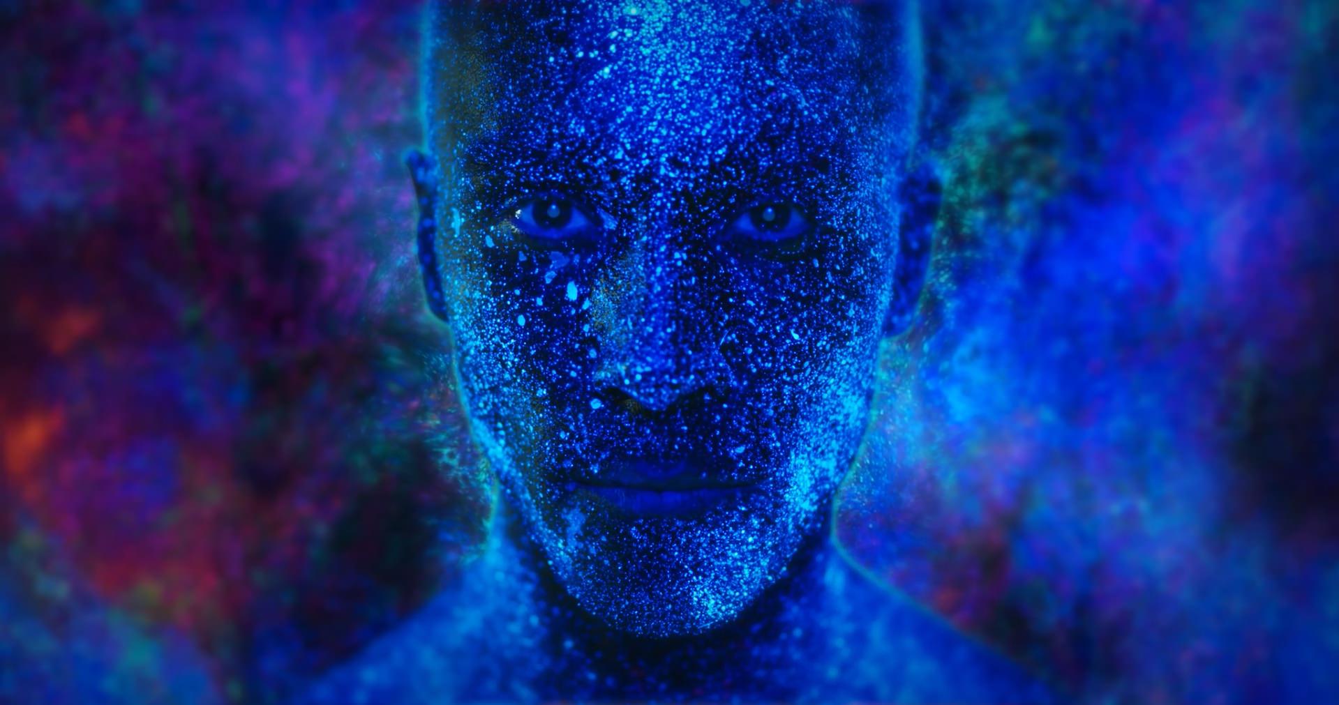 This image shows the face of man covered in blue and sparkly bodypaint surrounded by blue and purple glitter background