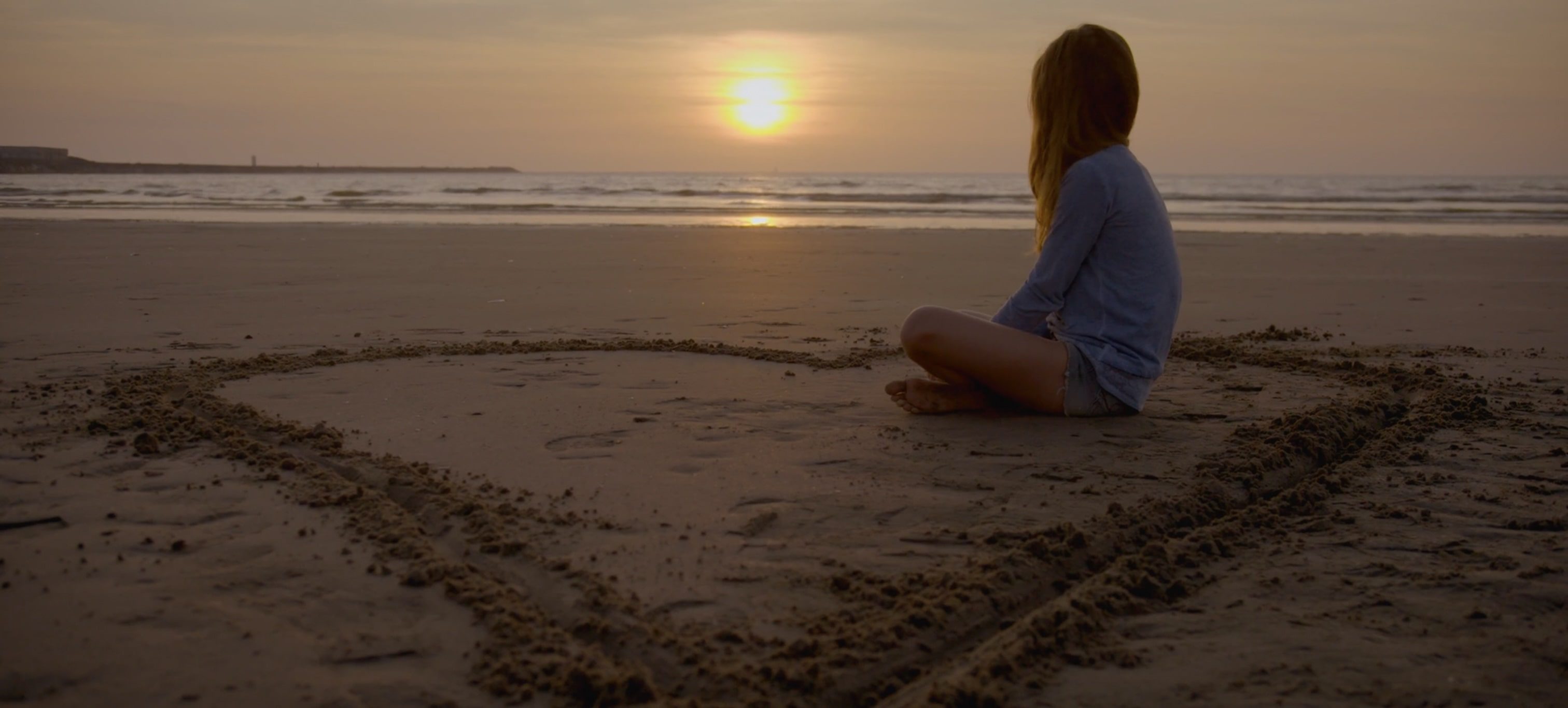 Young girl sitting on the beach at sunset in the middle of a large heart drawn in the sand